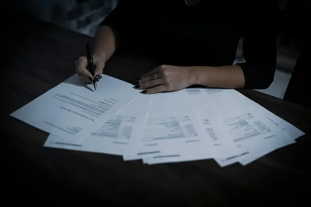 Des factures étalées sur une table sombre, une main tenant un stylo au-dessus.