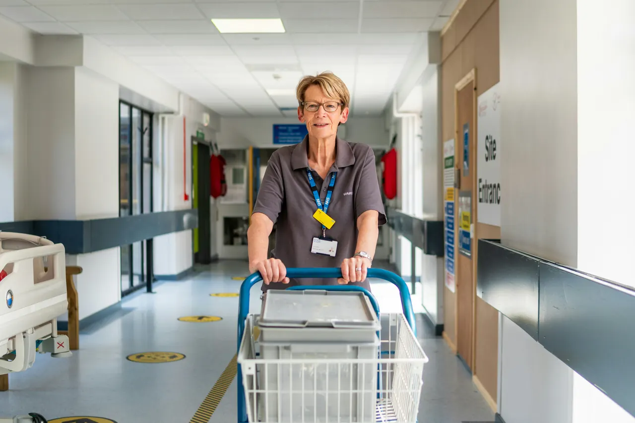 Une employée souriante qui pousse un chariot de fournitures dans un couloir d’hôpital.